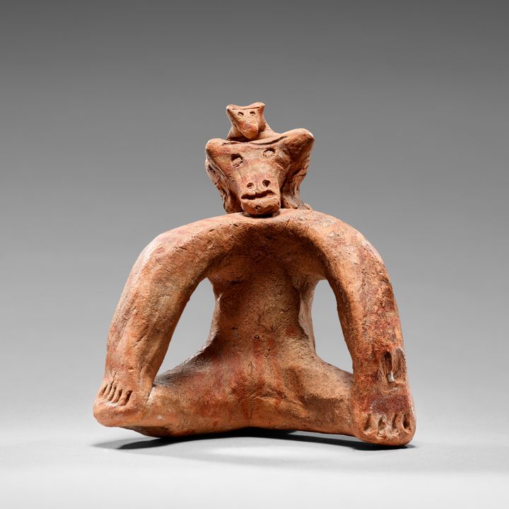 A red clay statuette shaped in the form of an ape sitting with its arms reaching out and down in an arc to the feet or knees. An upside-down triangular head features incised eyes, nose, and a mouth. A second tiny head perches at the top.