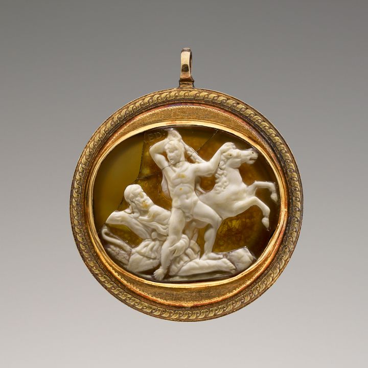 A honey-yellow cameo with a white upper layer showing two men and a horse in low relief. A nude man raises a club high over his head as he holds the rearing horse by its mane. The second man sits to the left of the scene and wraps one arm around the leg of the first. The cameo is set in a round gold frame.