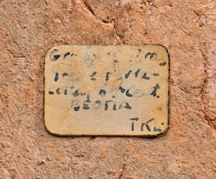 Close-up view of a sticker on a textured ceramic surface. The ink is faded which makes the five lines of text on it difficult to make out aside from the letter G at the top right, the phrase quote sixth cent end quote on the third line, and the word Beotia near the bottom.
