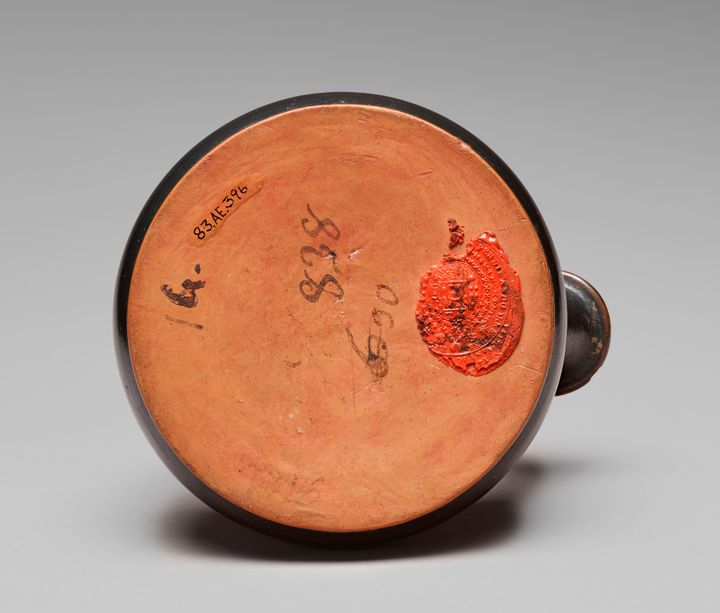 Underside of a ceramic vessel featuring several numbers and labels in black ink and a red wax seal. The seal is preserved partially to the right of the base, with an oval shield at the center encircled by rings of decorative marks and text.