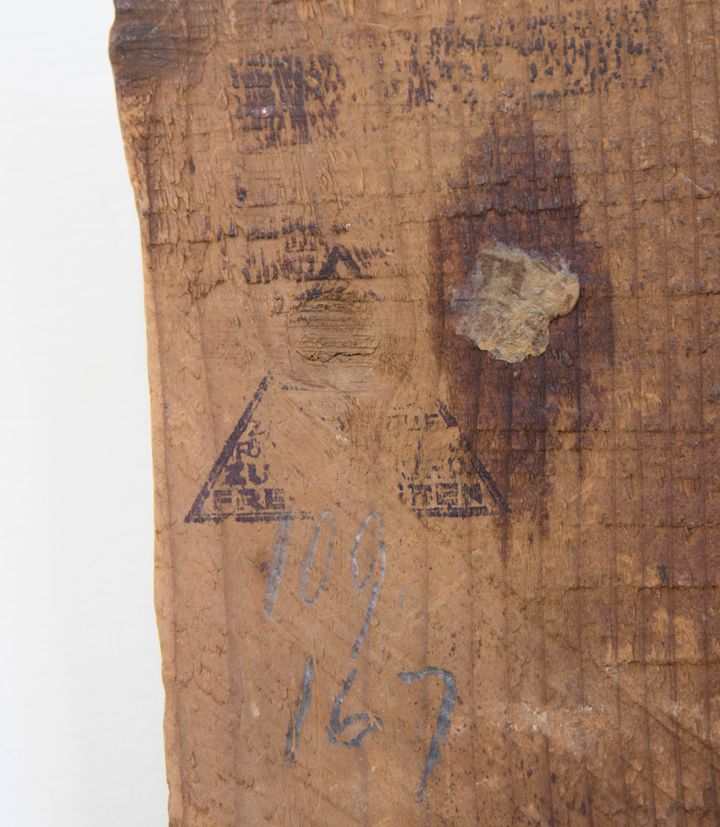 'A partial triangular stamp and two numbers written in pencil on a wooden panel. The center of the triangular stamp did not ink fully onto the wood grain, but a few letters are legible although they don''t make complete words.'