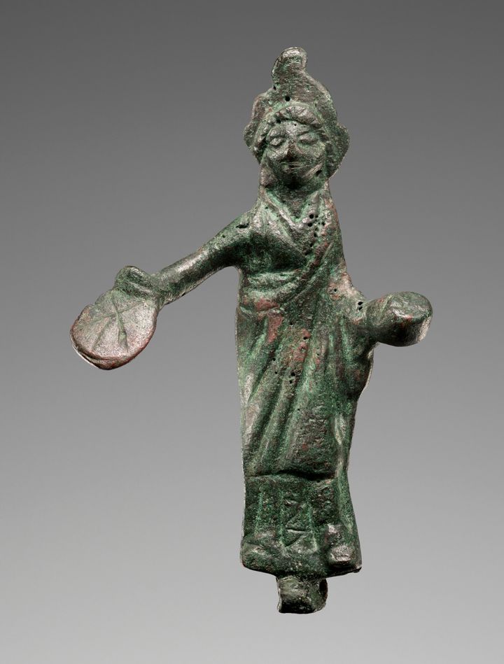 Bronze statuette showing a woman wearing a long garment and a headdress. She holds a round object out with her hand and a smaller dish-like object in her left. The pitted surface of the figure is streaked heavily with a green patina.