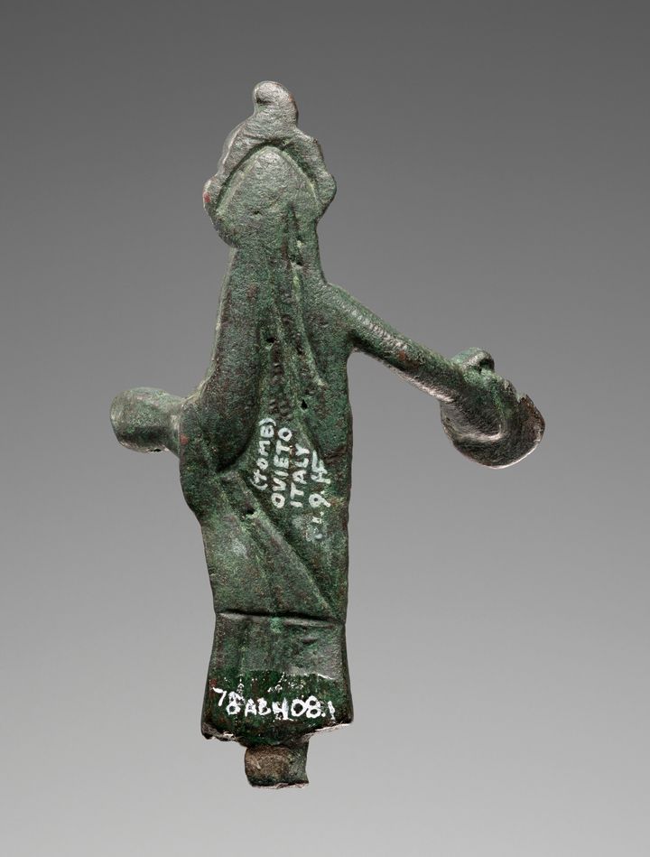 The back of the bronze statuette shown in Figure 20a. Two instances of white text are shown. Four lines oriented sideways in the center read quote Tomb Ovieto Italy 1.9.HF end quote, with the H and F conjoined. More white text along the bottom hem of the garment features an alphanumberic label.