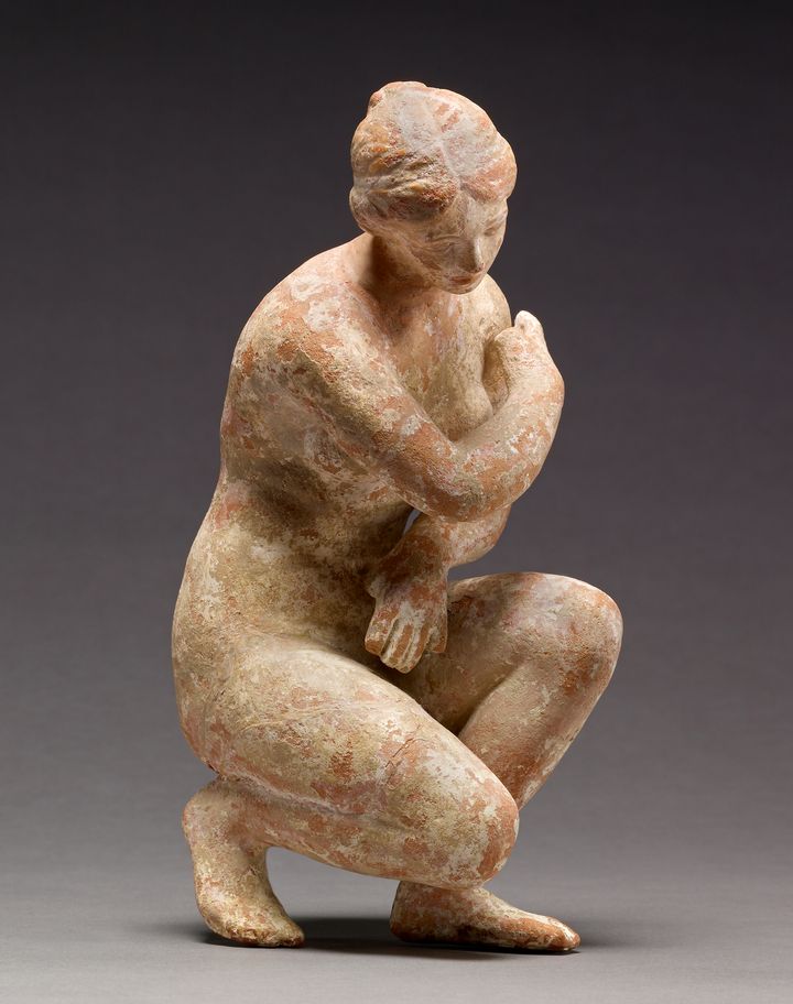 Terracotta statuette of a nude woman crouching and holding both arms across her chest. In this view, her body is angled to our right and her head tips to one side. Her hair is parted down the middle and pulled back.