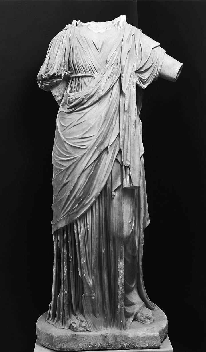 Black-and-white photograph of a marble statue of a woman wearing a long robe that falls in meticulous, dense pleats and creases. The head and most of her arms are missing. She stands with her weight on one foot and the other knee slightly bent, and one arm is lifted out to the side.