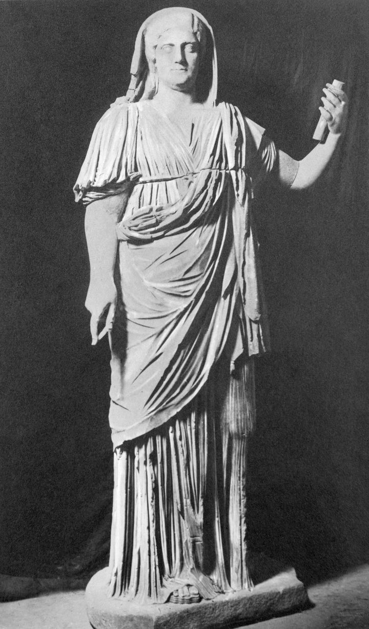 'Slightly blurry black-and-white photograph of the statue shown in Figure 23a but including the head and arms. The head is draped with a cloth, the right arm is extended by the side of the woman''s body, and the left holds up a cylindrical object, perhaps a fragment of an originally larger piece.'