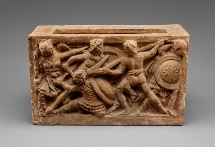View of one of the long sides of a terracotta trough-shaped urn. It shows a scene molded in relief with five people locked together in a struggle. A standing person prods a prone one with the end of a plowshare, which is as tall as him and has a hook reaching back at a sharp angle. Two of the people hold round shields, including the one on the ground.