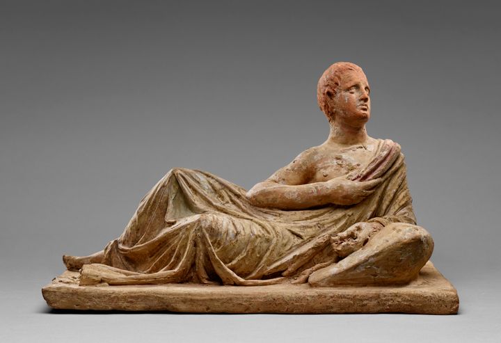Rectangular terracotta lid showing a reclining person with one elbow propped on a pillow. The person’s hips tip toward the front and the shoulders, to the right of the image, are squared to the viewer as the person looks up and to the right. They have short-cropped hair and wear a long toga that falls off one shoulder.