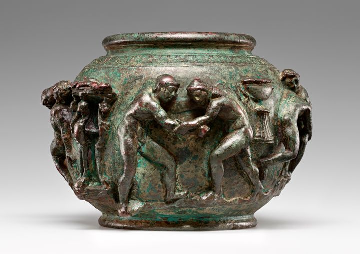 Bronze bowl with a rounded body and narrow neck and foot. Scenes in high relief decorate its sides. Two people appear wrestling on the side that faces the viewer. The surface gleams and has a green patina in areas.