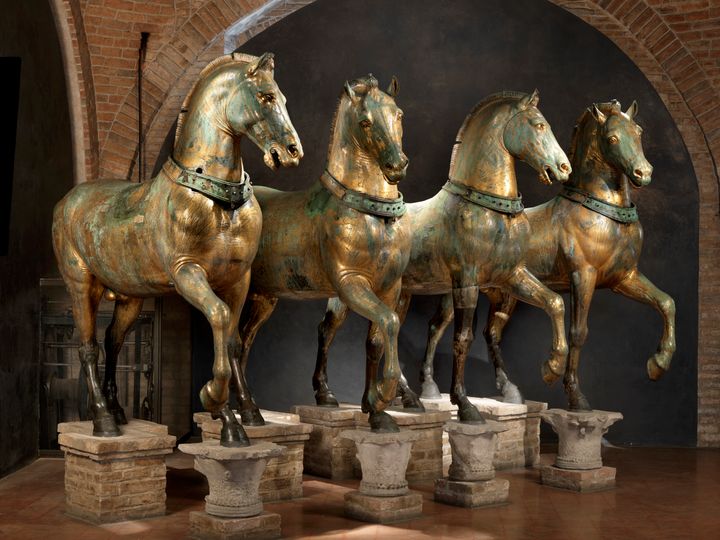 Row of four bronze fourses standing side by side on raised platforms. Their surfaces are muted gold streaked with pale green patina. Each horse stands on three legs and lifts a front leg. The front leg that remains on the ground rests on top of a capital. In pairs, the horses turn their heads slightly in toward each other and raise the same foreleg.