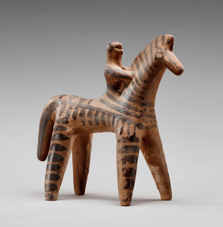 Terracotta statuette shaped into a simplified horse form with dark, parallel dashes. A person, smaller in scale than the animal, is on the horse’s back and clutches the mane. Their bodies meld together as the rider emerges from the back of the horse.