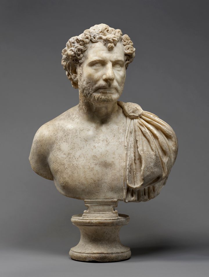 Marble bust showing the head, shoulders, and chest of a bearded man with short, curly hair. His lips are pulled tight and there are pouches on his cheeks under his eyes. His chest is bare except for a cloak pinned over his left shoulder. The bust rests on a round pedestal foot.