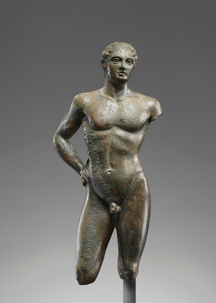 'One arm and the lower legs are missing from this bronze statuette of a nude young man. The preserved hand rests near his waist, and his hips jut to the right. He looks off to the right with deep-set eyes and his lips parted. Lettering appears down the left side of the statuette''s body from the armpit to where the knee cuts off.'