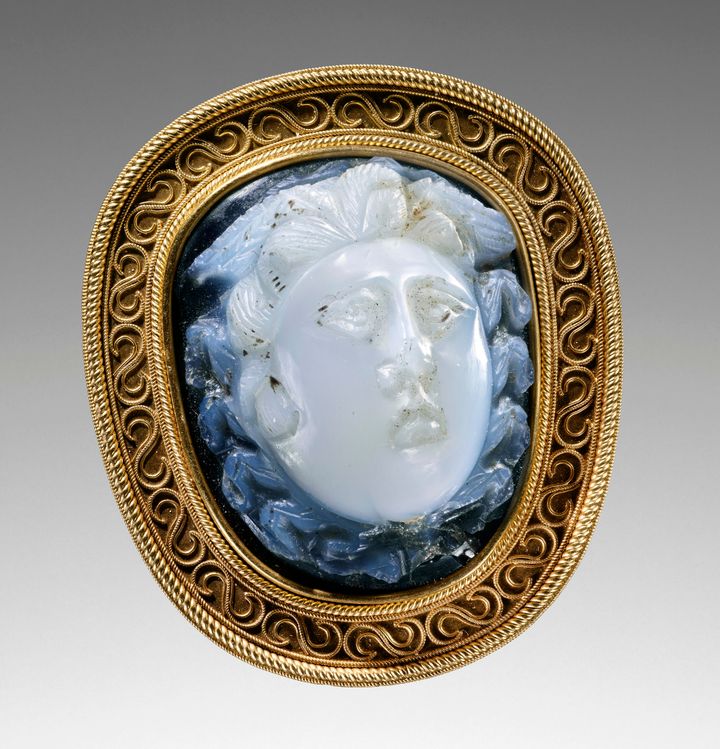 Cameo showing a person’s round face carved in low relief. The face appears iridescent white against a dark blue background. The eyes are close-set, and hair and snakes surround the head. The gold mount has repeating S-shapes set between two channels.