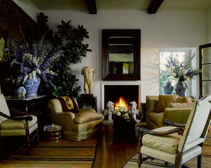 Marble sculptures of animals, a headless sculpted torso, and ceramic vessels are displayed in a living room among an overstuffed sofa and chair, armchairs, tables, and massive sprays of white and purple flowers in vases. A wood-framed mirror hangs over a lit fireplace.