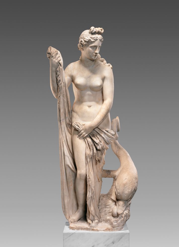 White marble statue of a mostly nude woman standing next to a stylized, downward-plunging fish or dolphin. Her body faces out, and she looks off to the right. She is depicted wrapping cloth across her hips with her left hand and holding the other end up in her right.