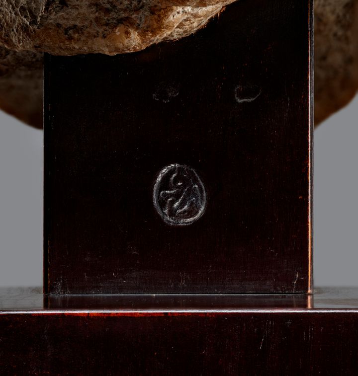 Close-up view of an oval stamp on the dark wood of a base, which supports a stone figure above.
