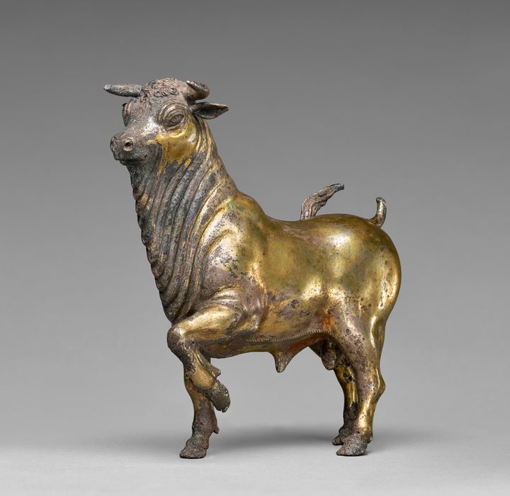 Statuette of a bull with short legs, a barrel-like body, a long, thick neck, and a partially lost upward flicking tail. Its left front leg is primly lifted, and folds cascade down the neck onto the chest. The eyes are slitted under arched brows. The surface is mostly golden though some areas are muted silver.