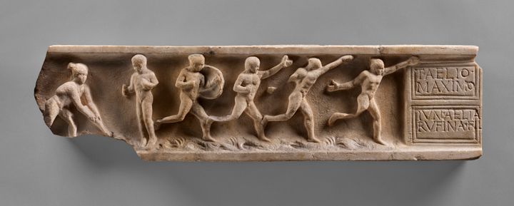 Tan-colored, rectangular marble relief showing two people standing facing each other and four people running off to the right. Three of the running people hold one hand high. A tablet at the right end of the fragment is incised with text in Latin.