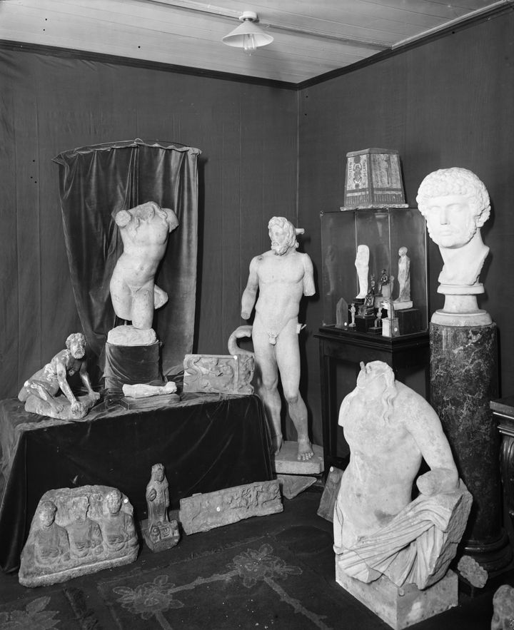 Black-and-white photograph of the corner of a room displaying Egyptian statuettes in a glass case, as well as fragmented stone reliefs, sculptures, and busts on tables and on the floor.