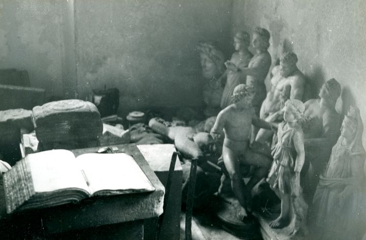 Black-and-white photograph of the corner of a room. The wall to the right is lined with statues on the floor. A thick book lies open on a table surface. Other objects fill the space.