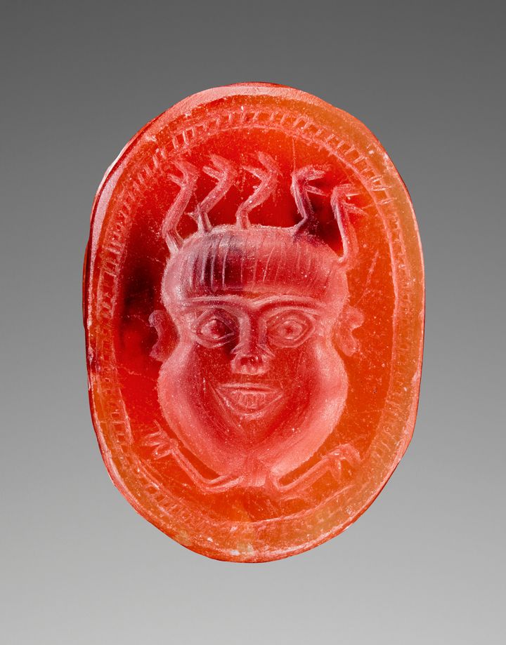 'Orange oval gem engraved with a round face with five snakes reaching from the top of the head and two extending out from the chin. The figure''s eyes are wide open and the mouth is pulled back in a toothy smile. The edge of the stone features linear incisions.'
