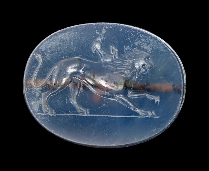 Blue oval gem engraved with a lion in profile facing the right and lifting one front paw as if mid-stride. It has the head, long neck, and front legs of a deer-like animal emerging from its shoulders.