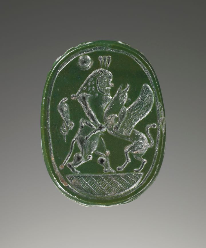 Green oval gem engraved with a standing person who is depicted fighting a griffin. The griffin features a lion’s body, wings, and the head of a bird of prey. The person has a long headdress and a rounded nose.