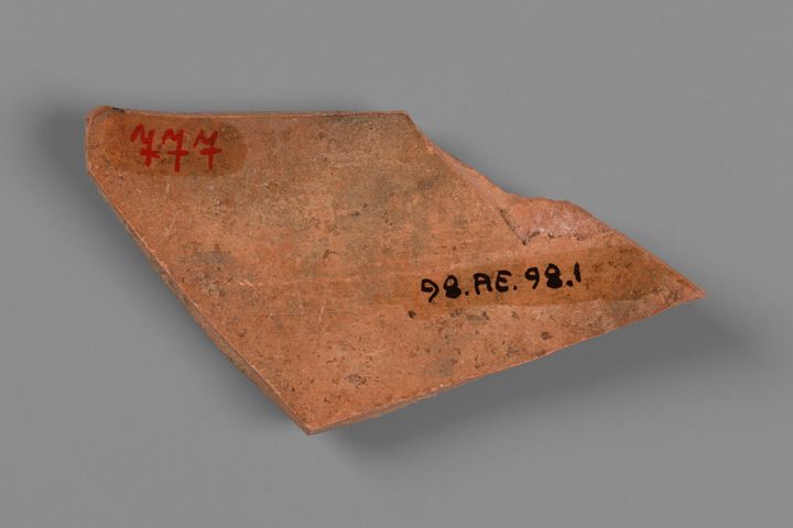 A roughly diamond-shaped pottery fragment with sharp edges and two numbers written on its surface. The number 777 is in red in the top left, and the label quote 98.AE.98.1 end quote is placed near the right tip in black.