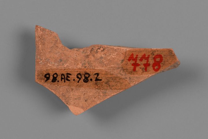 Terracotta fragment shaped in the form of a wide triangle with a spur at the top left. It has two numbers written on its surface: the label quote 98.AE.98.2 end quote is written in black to the left, and the number 778 is in red to the right.
