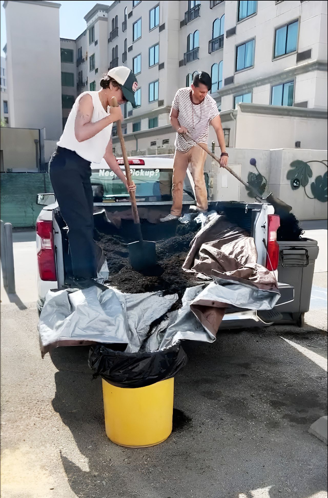 Two people stand in the back of a truck shoveling dirt into trash cans.