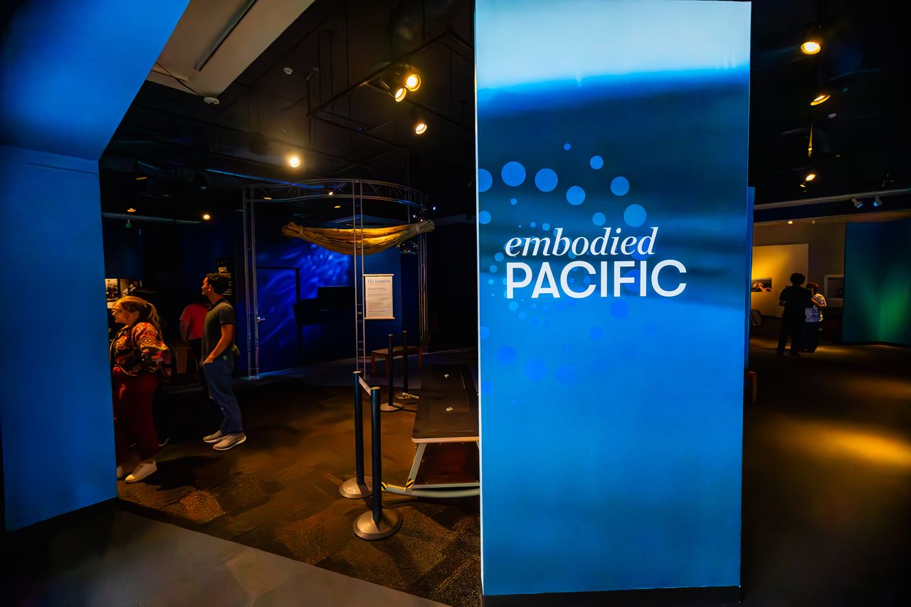 A large blue sign that says quote embodied Pacific end quote in a dark gallery with people looking at artwork.
