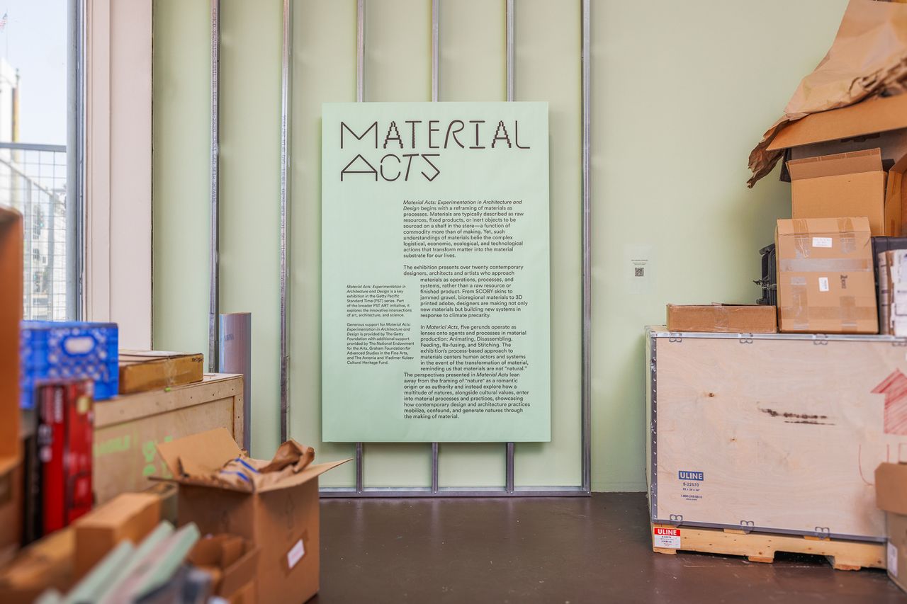 Green sign that says quote Material Acts end quote against a green wall with cardboard and storage boxes to the sides.