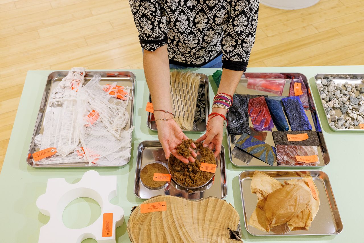 A hand holds up a grainy brown substance above a table of objects on silver display trays.