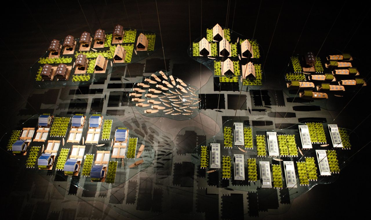 Architectural model-style installation featuring miniature buildings in varied styles and materials, arranged around a central leaf-shape.
