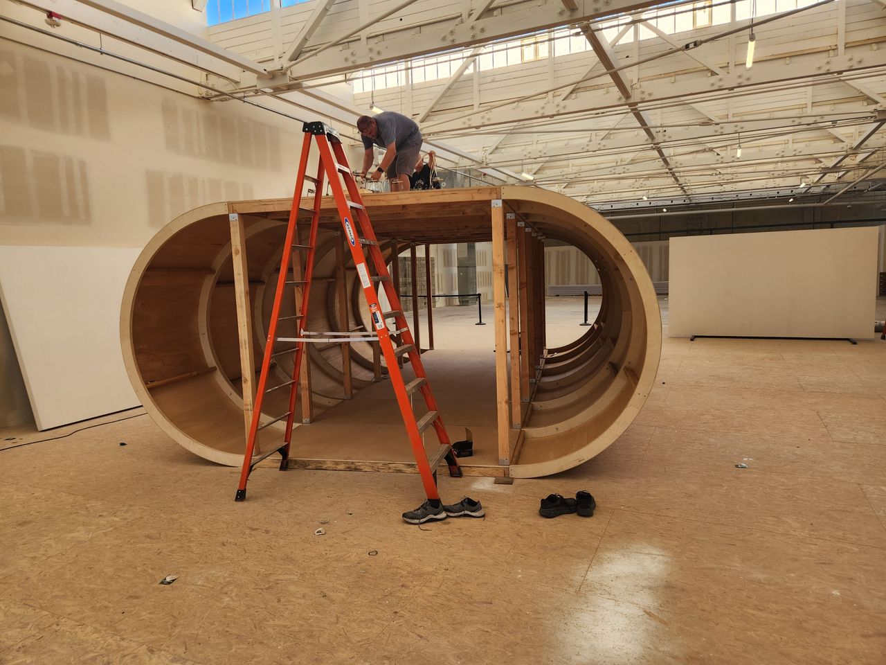 Red ladder and a person on top of a curved woodn structure in a gallery setting.