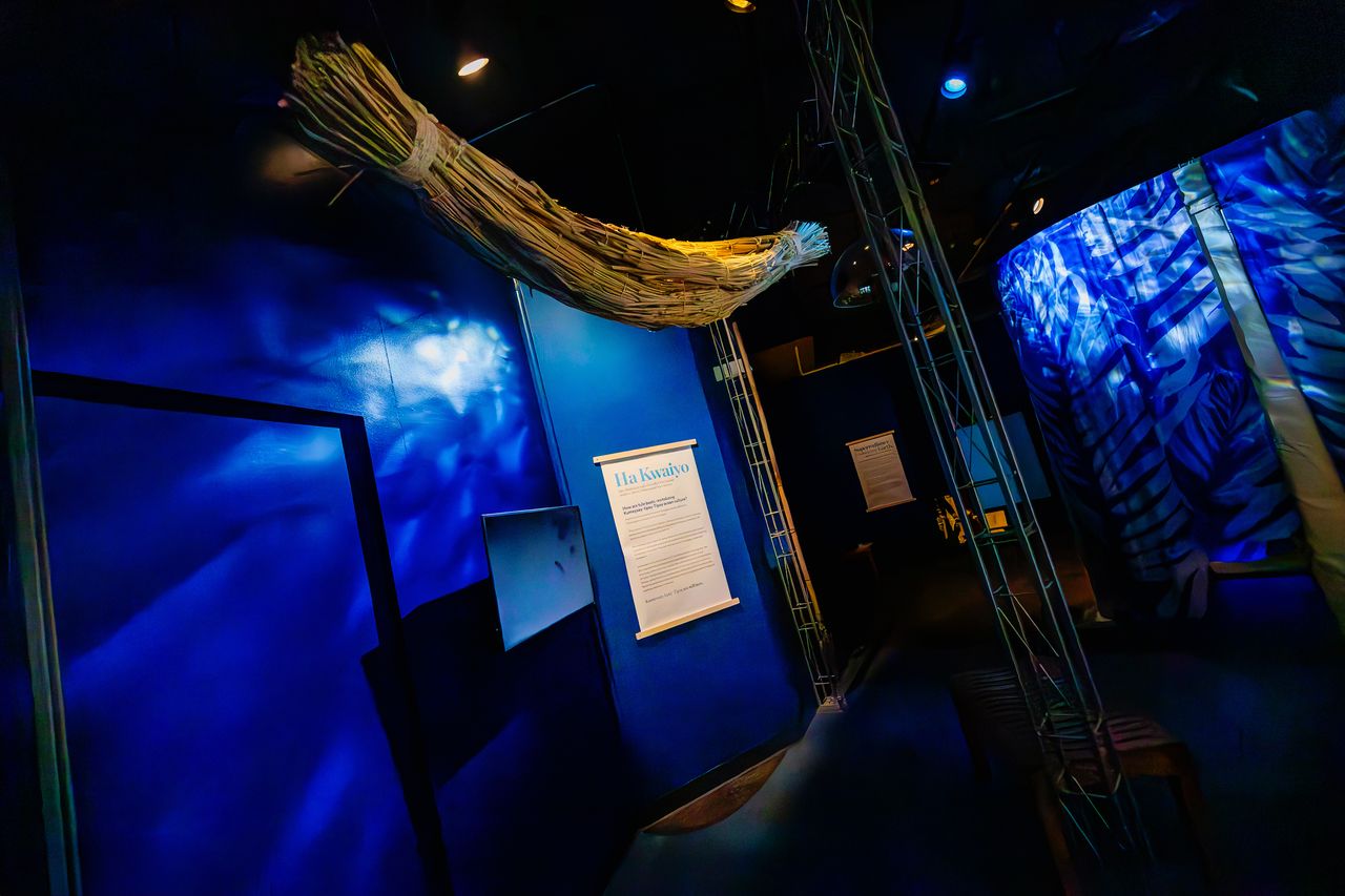Dark gallery setting with blue projections on the walls and a bundled stick sculpture handing from the ceiling and a sign that says quote Ha Kwaiyo end quote.