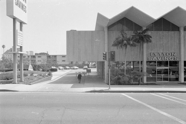 A black-and-white photo of a two-story building with a zigzag roofline. Above the door, a sign reads “Lytton Savings.”
