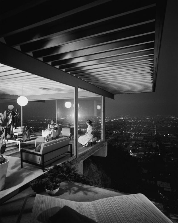 A black-and-white photo showing the exterior of a house at night, with floor-to-ceiling windows and a city view in the backdrop, as two women talk inside the living room.