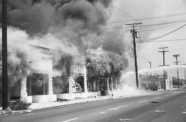 A black-and-white photo showing a street scene with several buildings on fire. Plumes of smoke rise into the sky.