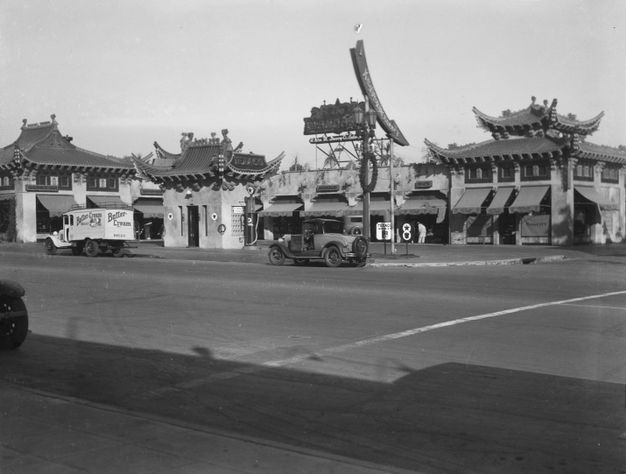 A black-and-white photo of a wide street with a series of buildings in the background. The buildings are built in architecture that resembles Chinese architecture, with slanted roofs that curve upwards at the tips.