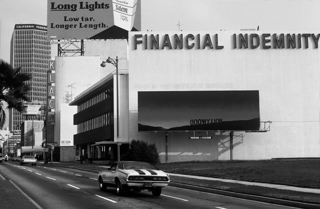 A black-and-white photo shows a city street with a line of buildings extending into the background. On the end of the closest building stands a billboard showing a sunset, and the back side of the Hollywood sign on the top edge of a mountain in shadow.