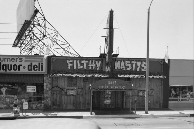A black-and-white photograph features a street with two building facades, the left of which reads liquor deli and the right which reads filthy nasty.