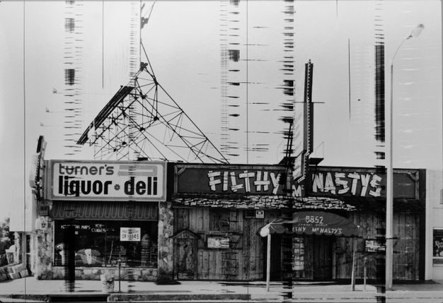 A black-and-white photograph features a street with two building facades, the left of which reads liquor deli and the right which reads filthy nasty. The photograph has been physically altered with scratches across its surface.