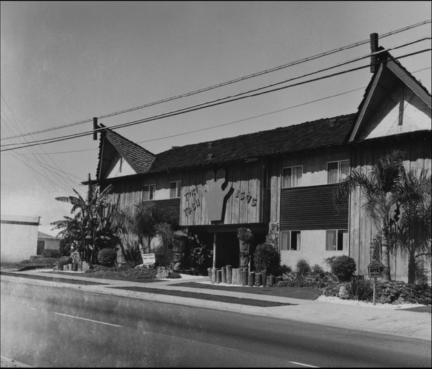 Two story apartment building with wood facade, shingled roof, and two decorative peaked sections. The building is named “Tiki Tabu” in large letters over the entrance.