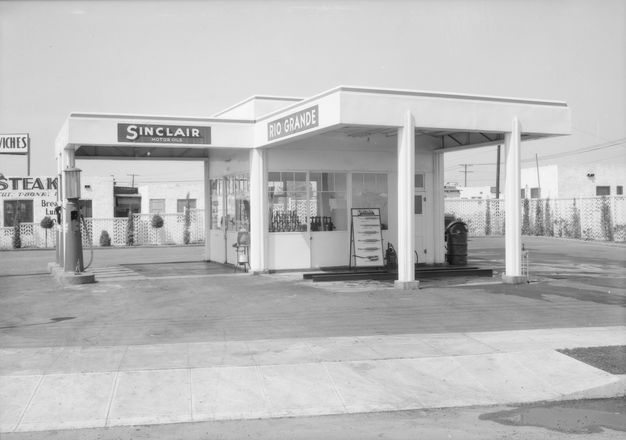A black-and-white photo features a gas station shot from the front. A sign on its façade reads “Sinclair.” There are no cars present.