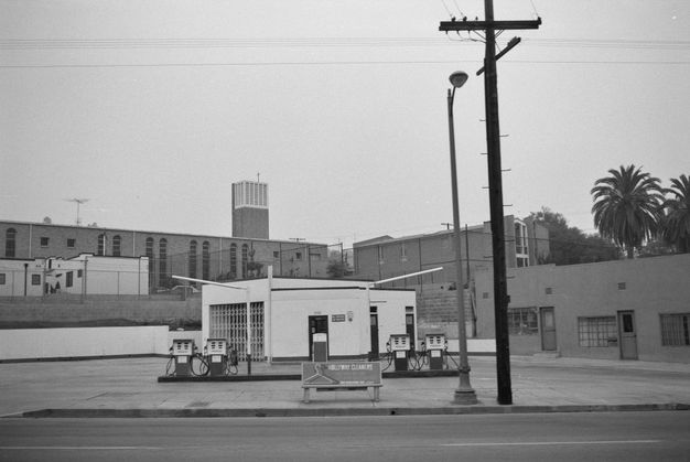 A small white gas station sits along an empty street. Numerous building surround it in the background.