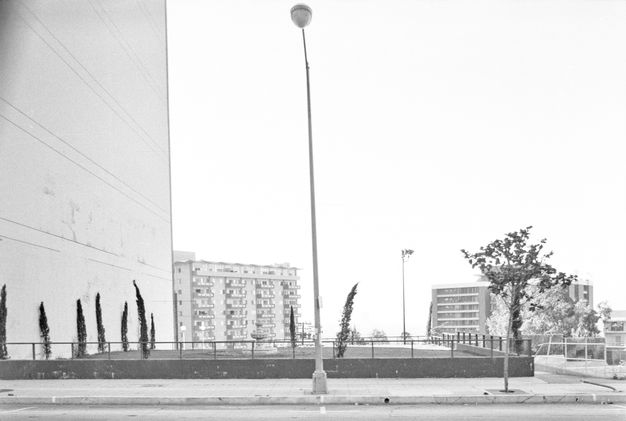 A black-and-white photo shows the side of a white building that extends above the frame on the left side of the image. In the background are two apartment buildings. In the center foreground stands a street lamp.