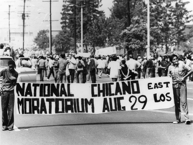A black-and-white photo featuring a group of people walking in the street in protest. Two people hold a long, horizontal banner that reads “national Chicano Moratorium June 29, East Los.”