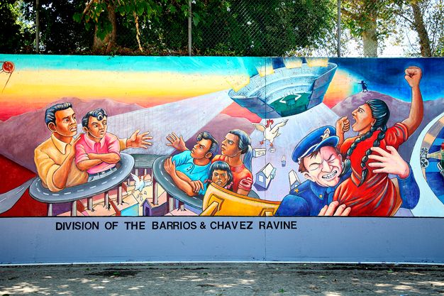 A mural depicting seven people, interlocking freeway designs, and a sunset. At right, a white police officer grabs a Latina woman and to the left are two Latinx families. Below, a caption reads “division of the barrios & chavez ravine.”